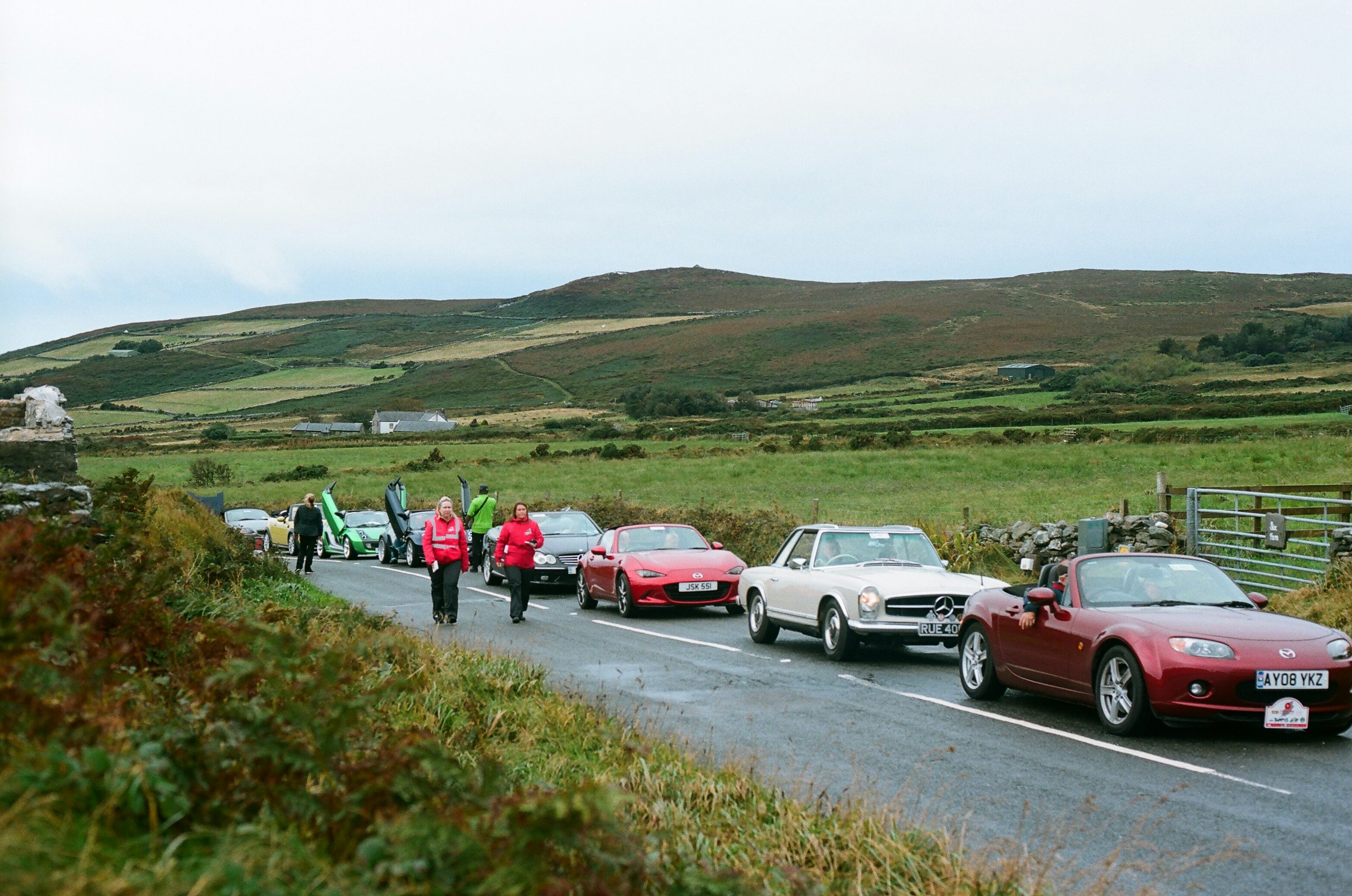 Cars and people on a rural road with hills.